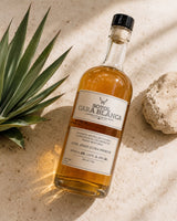 Licomar, Vinos y Licores Sotol Cara Blanca Añejo Ultra Premium 750ml bottle with amber liquid and black cap, on a sandy surface with agave plant.